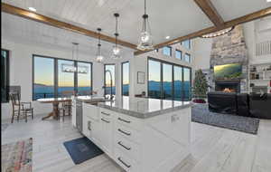 Kitchen with beam ceiling, white cabinetry, a stone fireplace, an island with sink, and pendant lighting