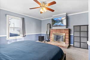 Remodeled bedroom featuring a brick fireplace, ceiling fan, crown molding and carpeted flooring