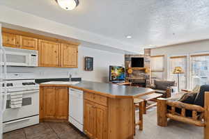 Kitchen with kitchen peninsula, a textured ceiling, white appliances, a stone fireplace, and light tile patterned flooring