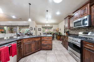 Kitchen with stainless steel appliances, dark brown cabinetry, dark stone countertops, decorative light fixtures, and light tile patterned floors