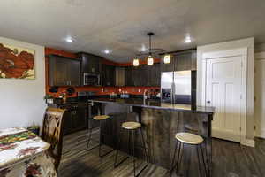 Kitchen with appliances with stainless steel finishes, dark brown cabinetry, a kitchen island with sink, dark wood-type flooring, and pendant lighting