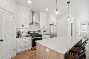 Kitchen featuring white cabinets, wall chimney range hood, light hardwood / wood-style flooring, appliances with stainless steel finishes, and a kitchen island