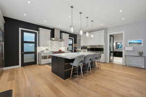 Kitchen featuring wall chimney range hood, hanging light fixtures, light wood-type flooring, a large island, and white cabinetry