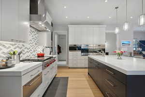 Kitchen featuring wall chimney range hood, pendant lighting, light hardwood / wood-style floors, a center island with sink, and white cabinets