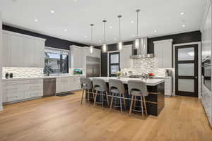 Kitchen featuring stainless steel appliances, wall chimney range hood, light hardwood / wood-style floors, decorative light fixtures, and a center island with sink