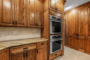 Kitchen with stainless steel double oven, light stone counters, decorative backsplash, brown cabinetry, and light tile patterned flooring