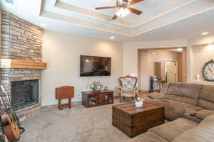 Carpeted living room with ceiling fan, a stone fireplace, and a tray ceiling