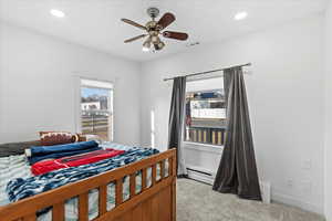 Carpeted bedroom featuring multiple windows, ceiling fan, and a baseboard radiator