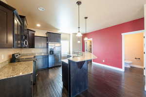 Kitchen featuring dark hardwood / wood-style flooring, light stone counters, stainless steel appliances, hanging light fixtures, and an island with sink