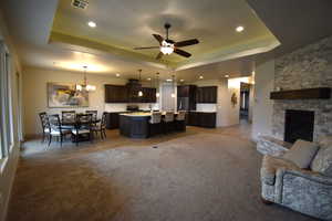 Carpeted living room featuring ceiling fan with notable chandelier, a stone fireplace, sink, and a tray ceiling