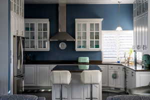 Kitchen with stainless steel refrigerator with ice dispenser, a kitchen breakfast bar, wall chimney range hood, pendant lighting, and white cabinets