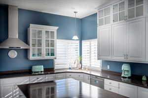 Kitchen featuring white cabinetry, dishwasher, sink, hanging light fixtures, and wall chimney range hood