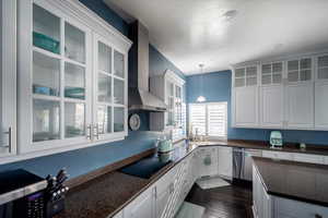 Kitchen with black electric stovetop, stainless steel dishwasher, wall chimney exhaust hood, decorative light fixtures, and white cabinets