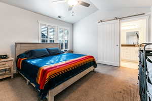 Carpeted bedroom with a barn door, ensuite bathroom, vaulted ceiling, and ceiling fan