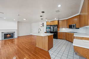 Kitchen with stainless steel appliances, a premium fireplace, light hardwood / wood-style floors, decorative light fixtures, and a center island with sink