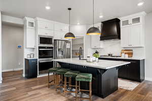 Kitchen featuring pendant lighting, custom exhaust hood, white cabinets, an island with sink, and appliances with stainless steel finishes