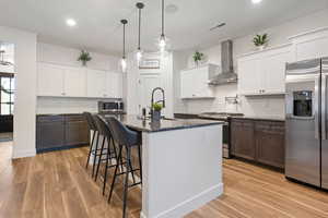 Kitchen featuring wall chimney exhaust hood, stainless steel appliances, a kitchen island with sink, light hardwood / wood-style flooring, and white cabinets