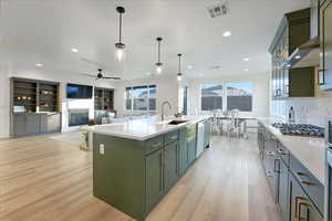 Kitchen featuring sink, wall chimney range hood, an island with sink, decorative light fixtures, and light wood-type flooring