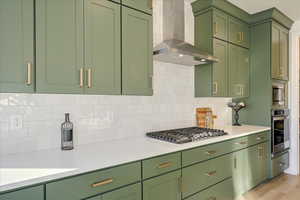 Kitchen featuring wall chimney exhaust hood, stainless steel appliances, green cabinets, backsplash, and light hardwood / wood-style floors