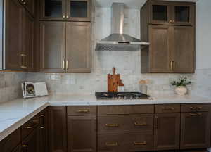 Kitchen with stainless steel gas stovetop, decorative backsplash, light stone counters, and wall chimney range hood