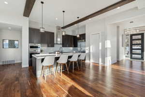 Kitchen with dark brown cabinetry, wall chimney range hood, dark hardwood / wood-style flooring, beamed ceiling, and a kitchen island with sink