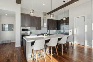 Kitchen featuring a kitchen island with sink, wall chimney exhaust hood, dark hardwood / wood-style floors, appliances with stainless steel finishes, and dark brown cabinets