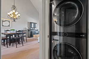 Clothes washing area featuring carpet floors, stacked washer and dryer, and a notable chandelier