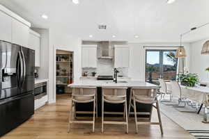 Kitchen featuring hanging light fixtures, stainless steel appliances, wall chimney range hood, light hardwood / wood-style floors, and white cabinets