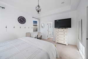 Bedroom featuring a raised ceiling, light carpet, ensuite bathroom, and a notable chandelier