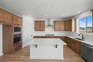 Kitchen with wall chimney range hood, sink, a kitchen island, appliances with stainless steel finishes, and light hardwood / wood-style floors