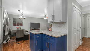 Kitchen with blue cabinetry, tasteful backsplash, stainless steel fridge, wood-type flooring, and ceiling fan with notable chandelier