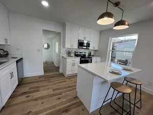 Kitchen with stainless steel appliances, decorative light fixtures, hardwood / wood-style flooring, white cabinetry, and a breakfast bar area