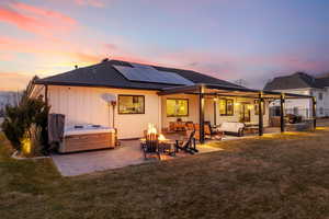 Back house at dusk featuring a yard, solar panels, a hot tub, an outdoor living space with a fire pit, and a patio