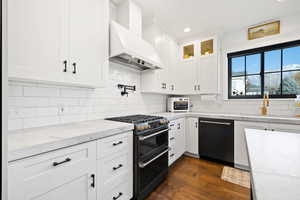 Kitchen with backsplash, premium range hood, dark wood-type flooring, appliances with stainless steel finishes, and white cabinetry