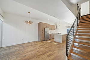 Kitchen featuring light wood-type flooring, stainless steel appliances, wall chimney range hood, decorative light fixtures, and a chandelier