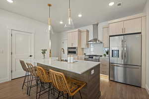 Kitchen with appliances with stainless steel finishes, light brown cabinetry, wall chimney exhaust hood, sink, and an island with sink