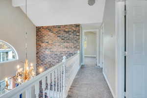 Hallway featuring lofted ceiling with beams, light colored carpet, brick wall, and an inviting chandelier