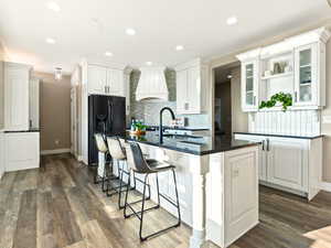 Kitchen with black fridge, custom exhaust hood, dark wood-type flooring, white cabinets, and an island with sink