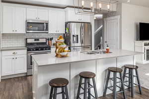 Kitchen with dark wood-type flooring, an island with sink, appliances with stainless steel finishes, decorative light fixtures, and white cabinetry