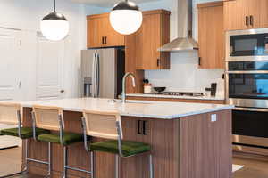 Kitchen featuring sink, wall chimney range hood, wood-type flooring, a center island with sink, and appliances with stainless steel finishes