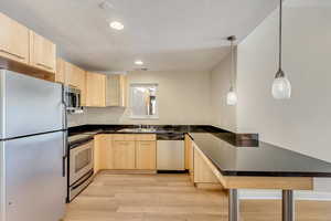 Kitchen featuring light brown cabinets, hanging light fixtures, stainless steel appliances, light hardwood / wood-style flooring, and kitchen peninsula