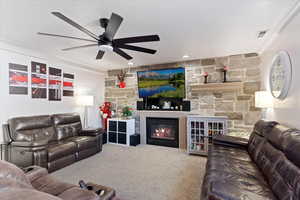 Living room with carpet, a stone fireplace, ceiling fan, and crown molding