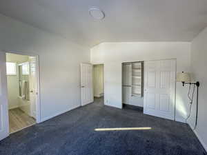 Unfurnished bedroom featuring ensuite bath, a closet, lofted ceiling, and dark colored carpet