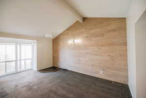 Unfurnished room featuring dark colored carpet, vaulted ceiling with beams, and wood walls