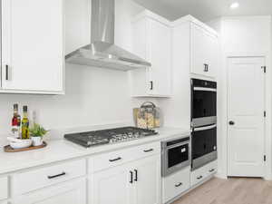 Kitchen featuring wall chimney range hood, light wood-type flooring, appliances with stainless steel finishes, light stone counters, and white cabinetry