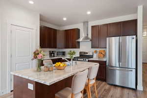 Kitchen featuring a center island with sink, appliances with stainless steel finishes, light wood-type flooring, a kitchen bar, and wall chimney range hood