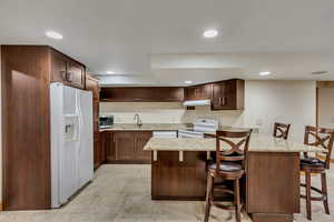 Kitchen featuring sink, light stone counters, kitchen peninsula, white appliances, and a breakfast bar