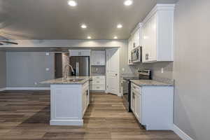 Kitchen with light stone counters, stainless steel appliances, dark wood-type flooring, white cabinets, and an island with sink