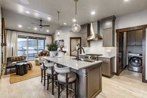 Kitchen featuring wall chimney range hood, washer / clothes dryer, decorative light fixtures, a kitchen island with sink, and light wood-type flooring