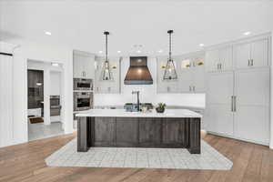 Kitchen featuring a center island with sink, white cabinetry, light wood-type flooring, and premium range hood